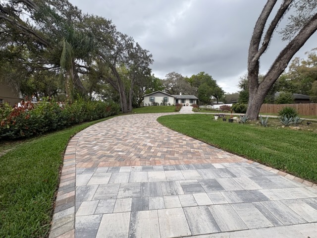 Curving paver driveway leading to a house framed by mature trees and fresh lawn